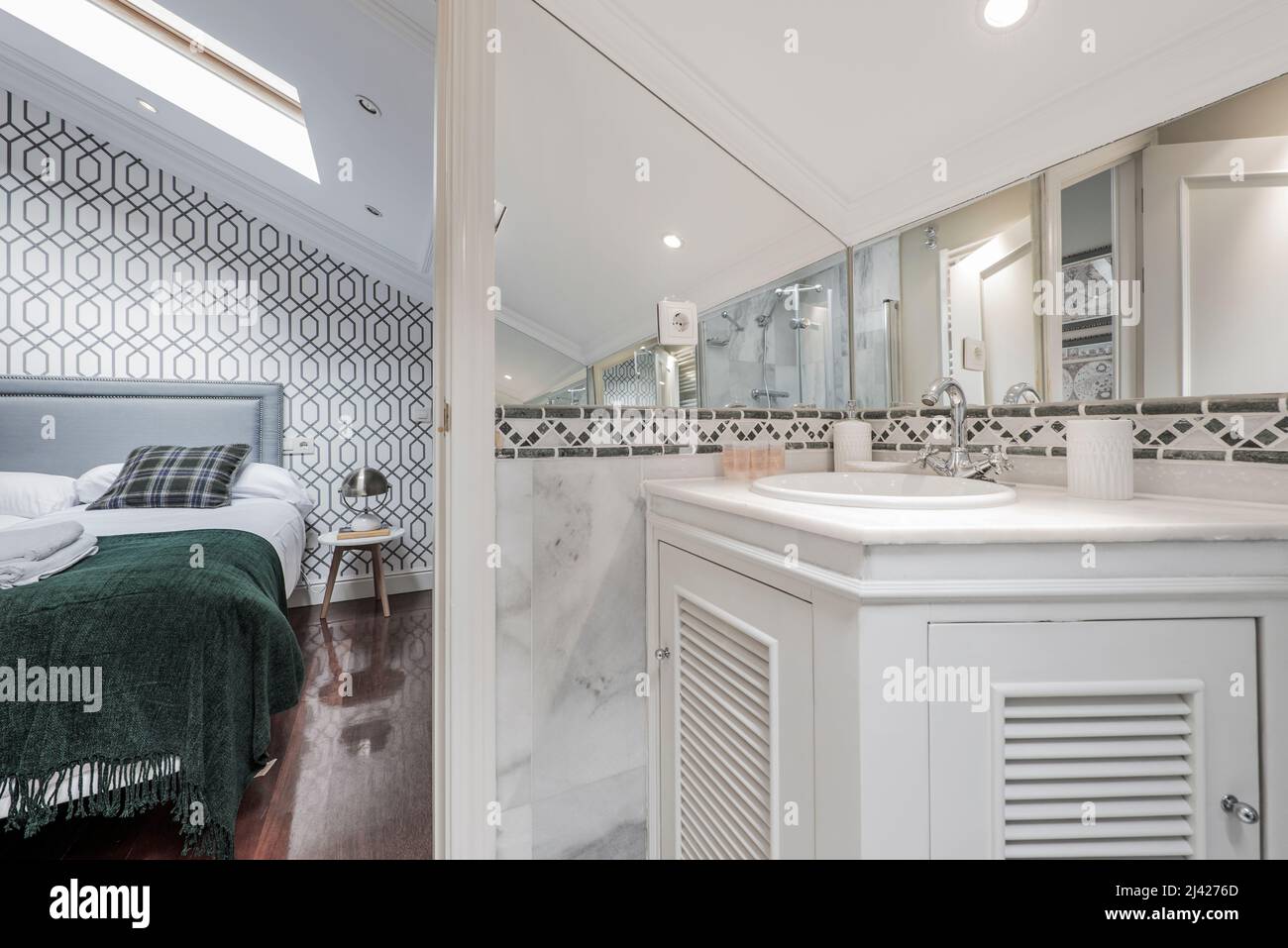 Bedroom with double bed and en-suite bathroom with white wooden furniture, white marble walls and mirror in a room with sloping ceilings Stock Photo