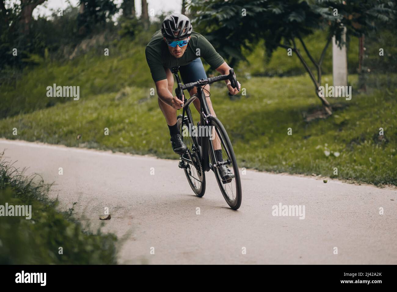 Caucasian strong man in safety helmet and mirrored glasses dynamically riding bicycle on road among green park. Morning time for exercises on fresh ai Stock Photo