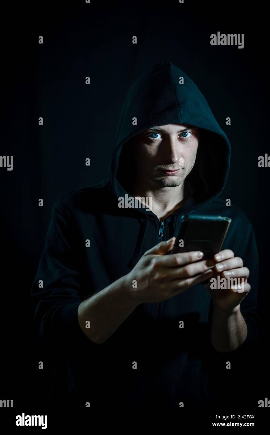 Portrait of a young hooded man who is using a smarphone and looking at the camera as if he was disturbed Stock Photo
