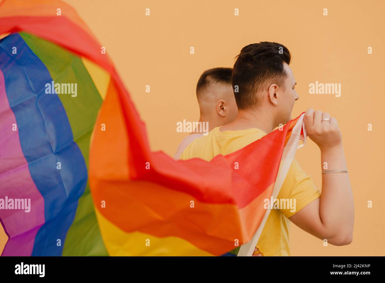 Young gay couple holding rainbow flag while standing over yellow wall ...