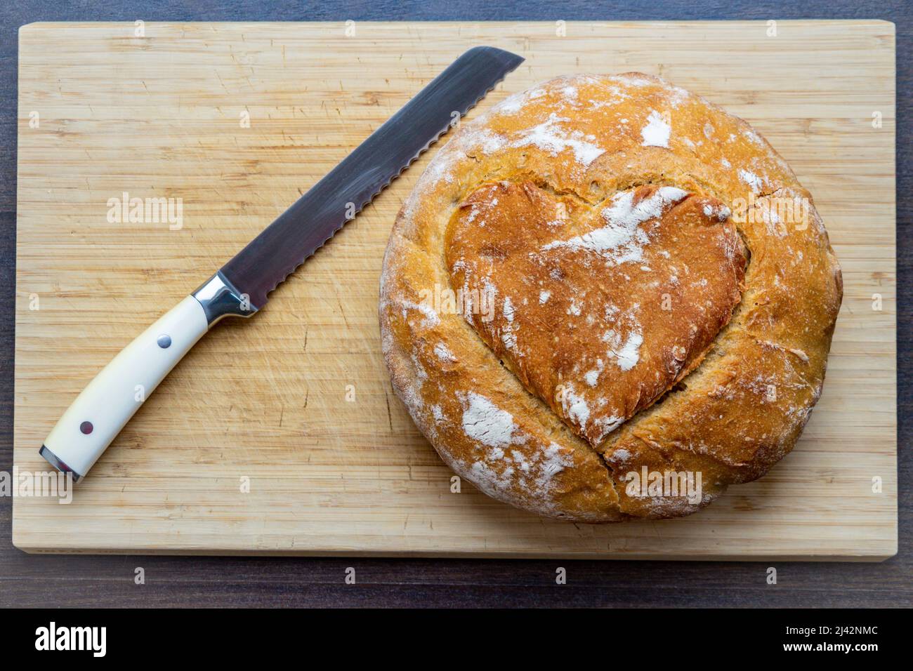 A freshly baked bread with a heart carved into the crust lies on a wooden slate beside a bread knife Stock Photo