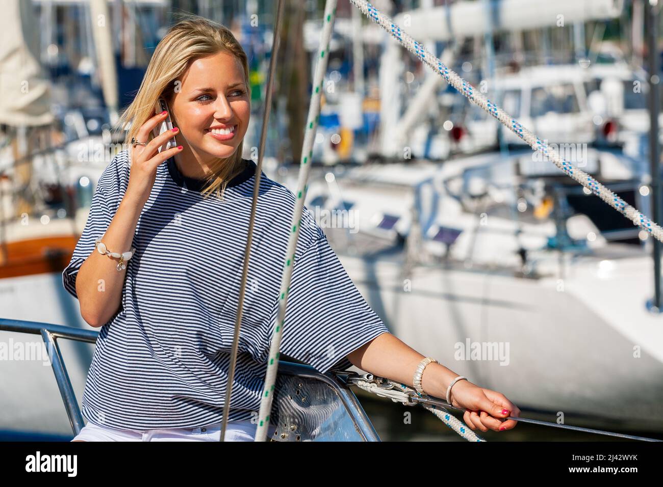 Beautiful young woman blonde girl sitting on the deck of a boat yacht talking on a mobile cell phone in summer sunshine Stock Photo