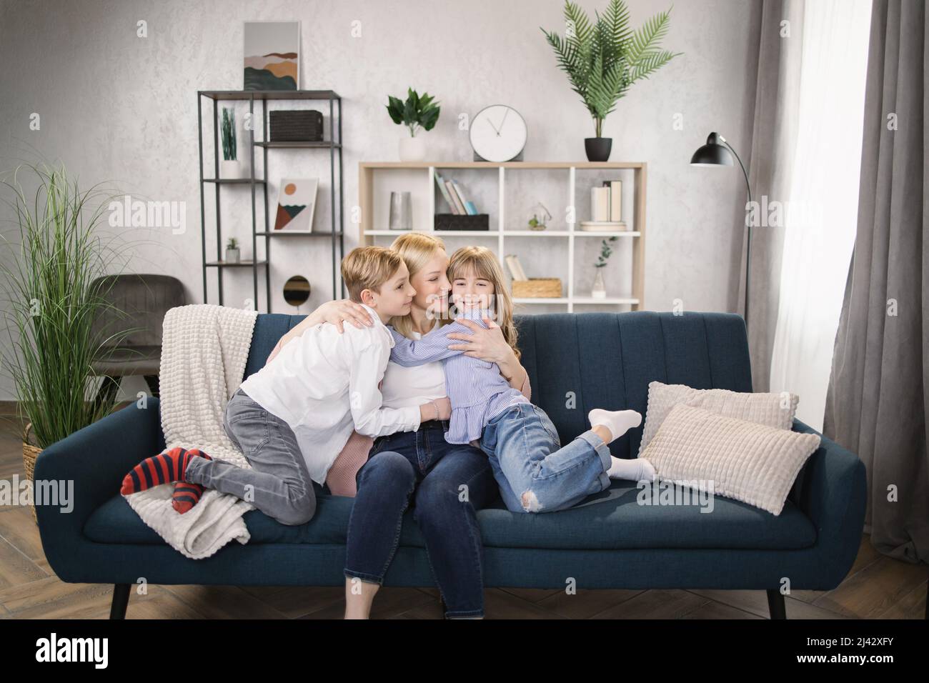 Happy little kids sit on couch hug cuddle excited young mom or nanny show  love and care, overjoyed small children relax with mother embrace share  sweet tender family moment in living room