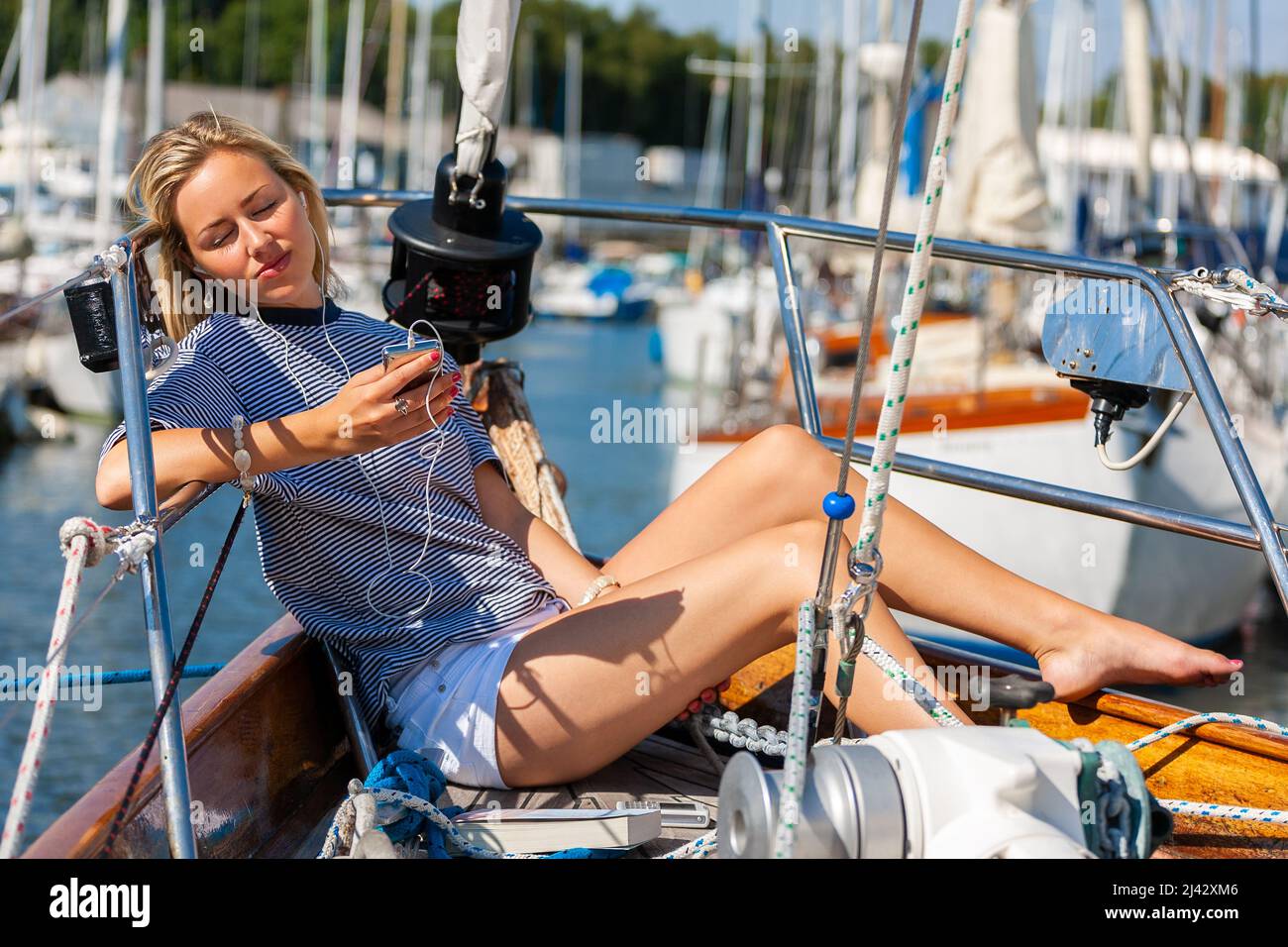 Beautiful young woman blonde girl eyes closed, relaxing with a book, sitting on the deck of a boat yacht listening to music on earphones and a mobile Stock Photo