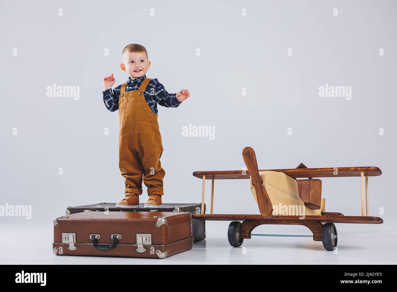 A cheerful child in a brown overalls is standing on a suitcase. Little traveler with a suitcase