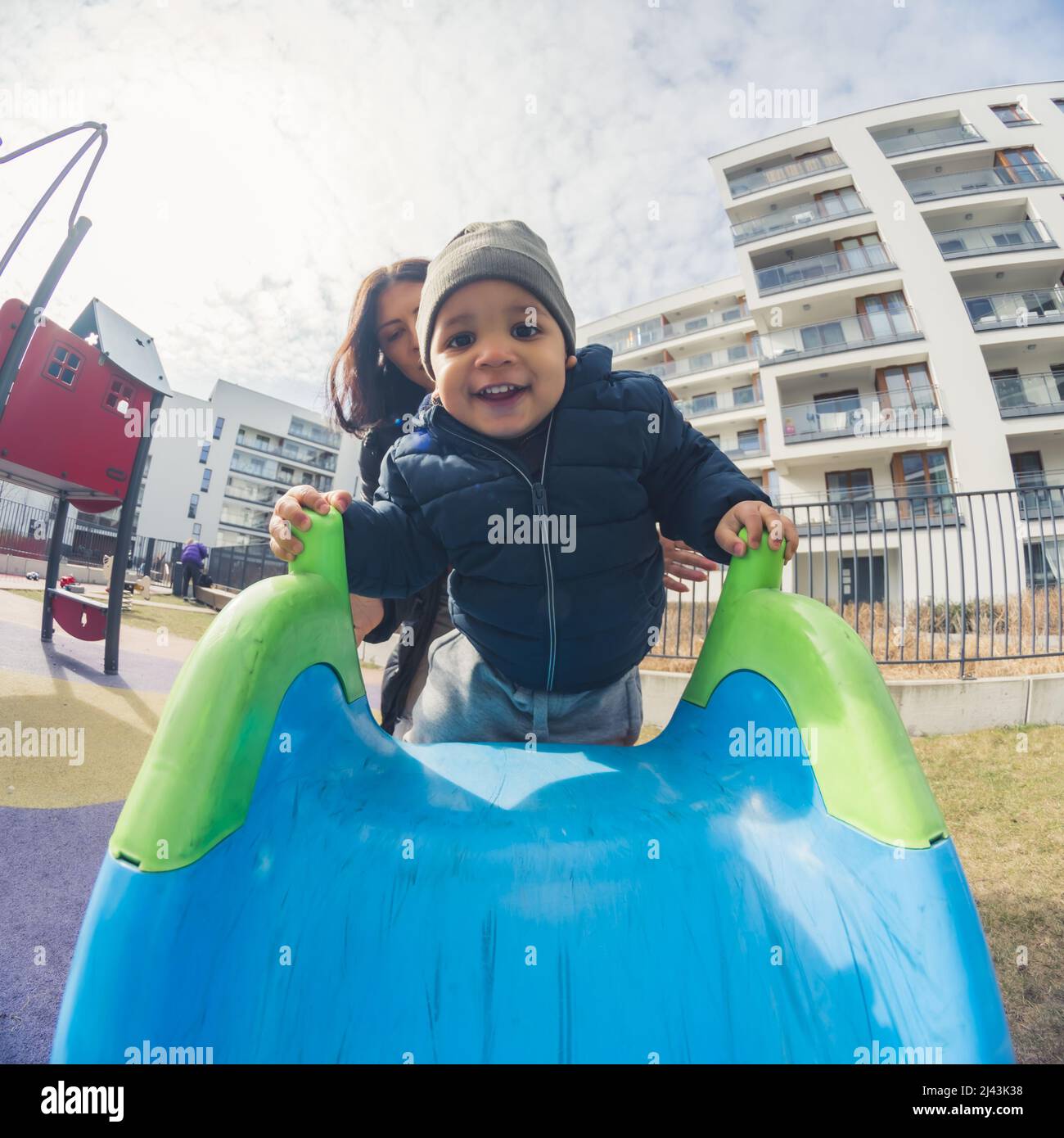 happy African American little boy with a grey hat in front of his mother climbing on a blue slide in the park buildings background cowboy shot. High quality photo Stock Photo