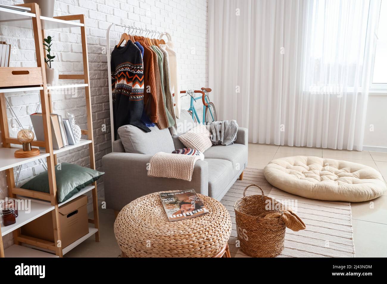 Sofa, shelf unit and rack with sweaters near white brick wall in living ...