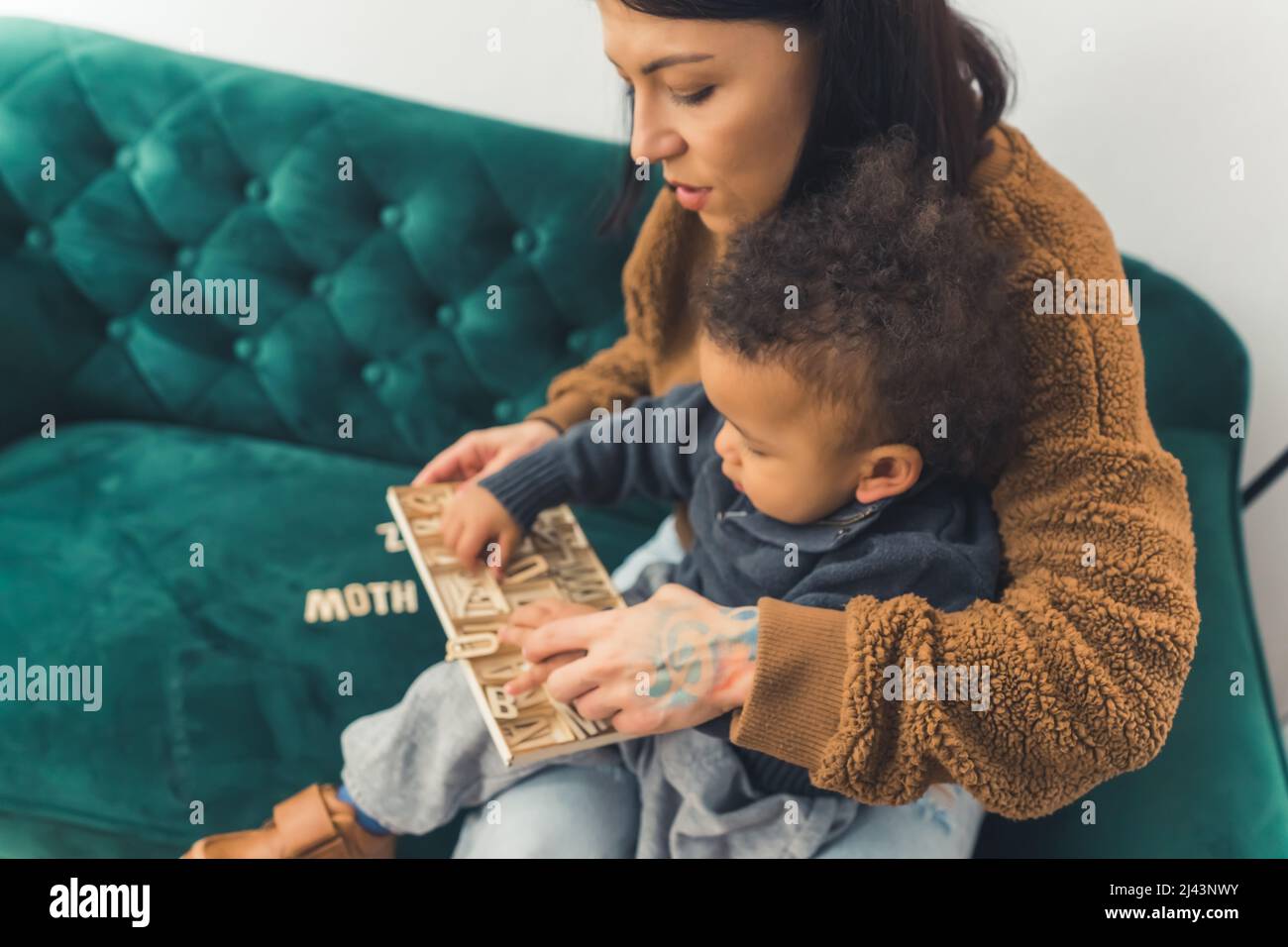 lovely curled little boy sitting in his mother's lap who is sitting on the green sofa and looking at something with his mother medium full shot indoor copy space. High quality photo Stock Photo