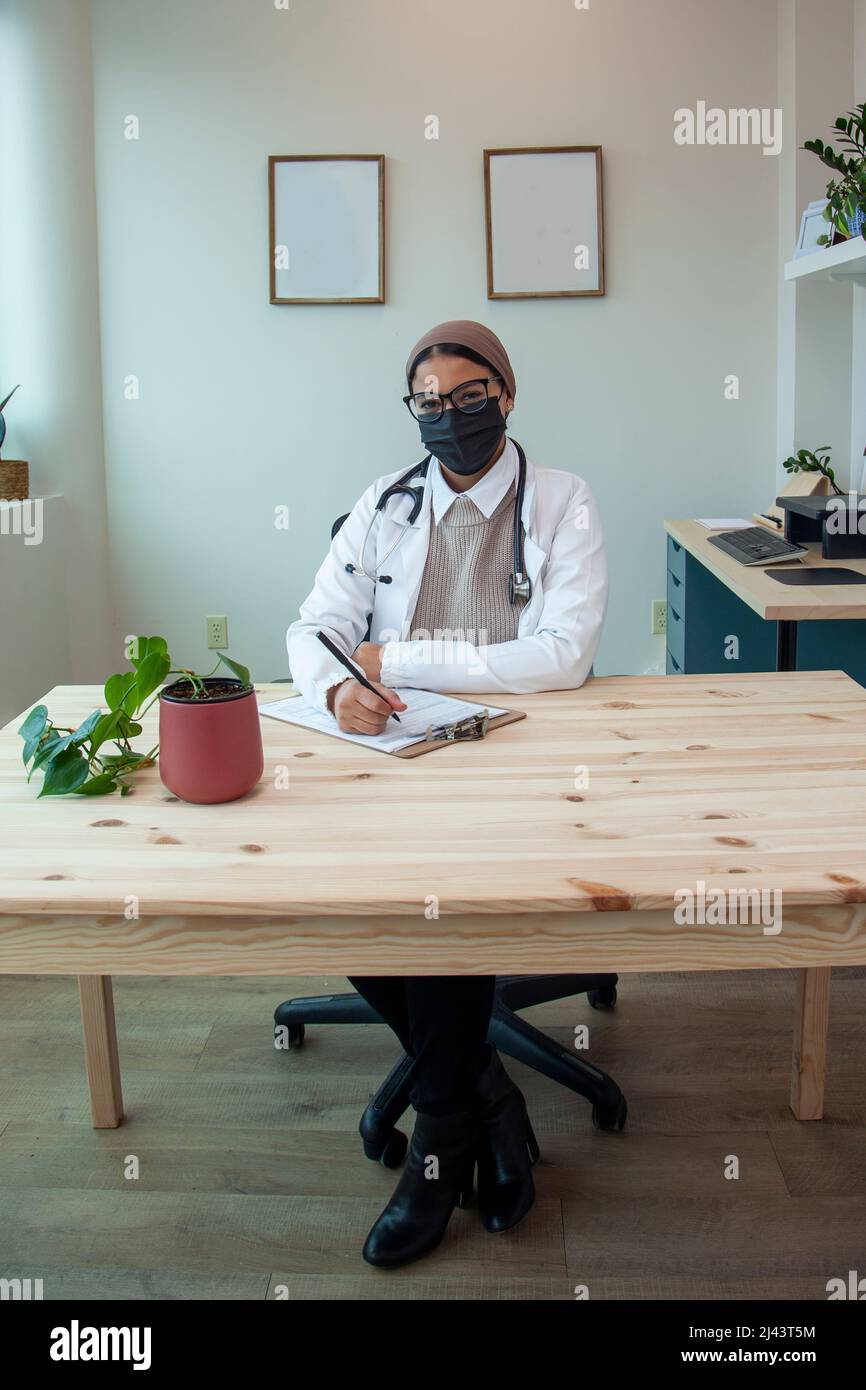 a doctor in a mask sits working in her office smiling and doing paperwork Stock Photo