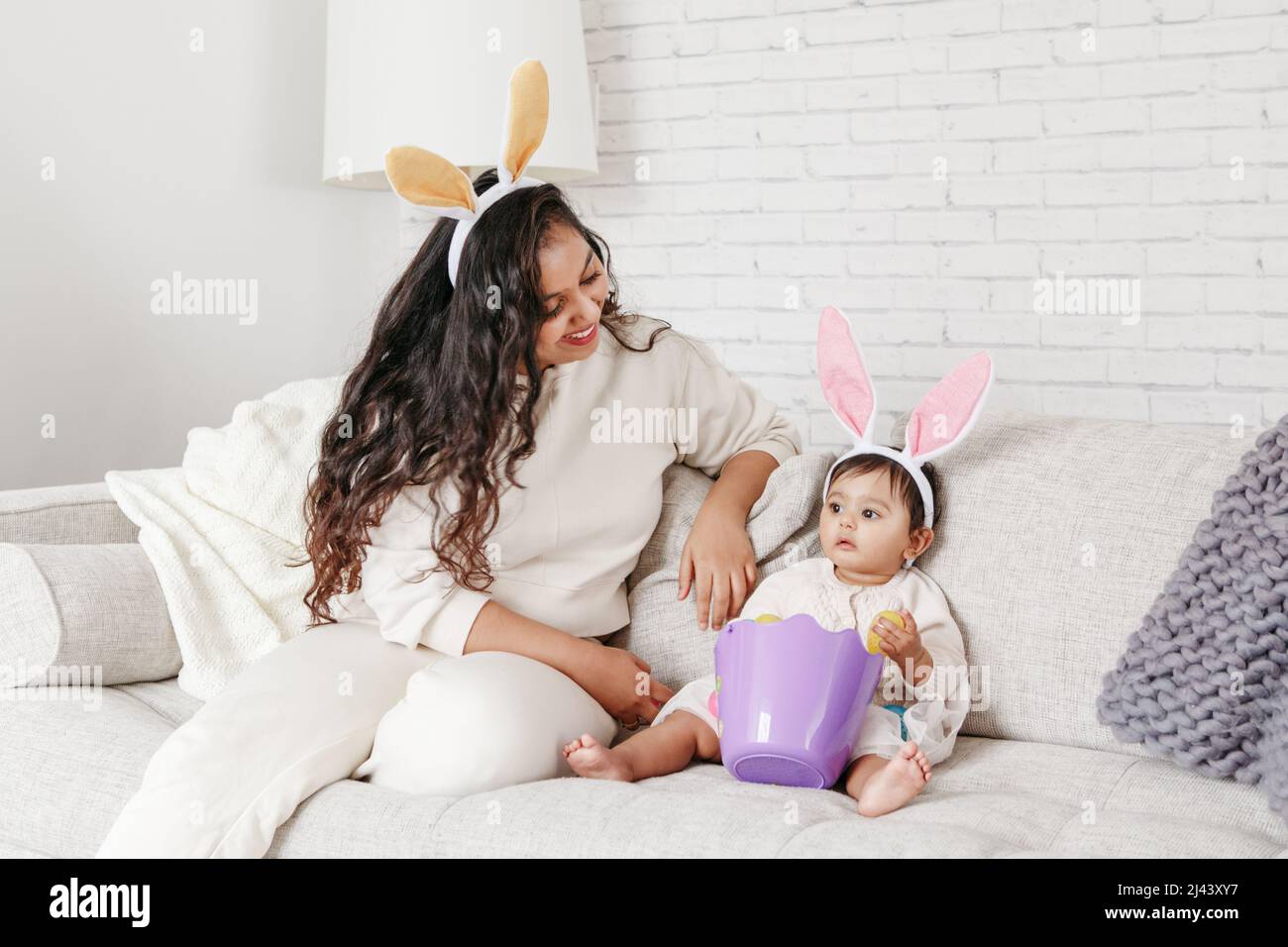 Indian mother with baby girl daughter in bunny ears playing toy eggs in basket celebrating Easter. Stock Photo