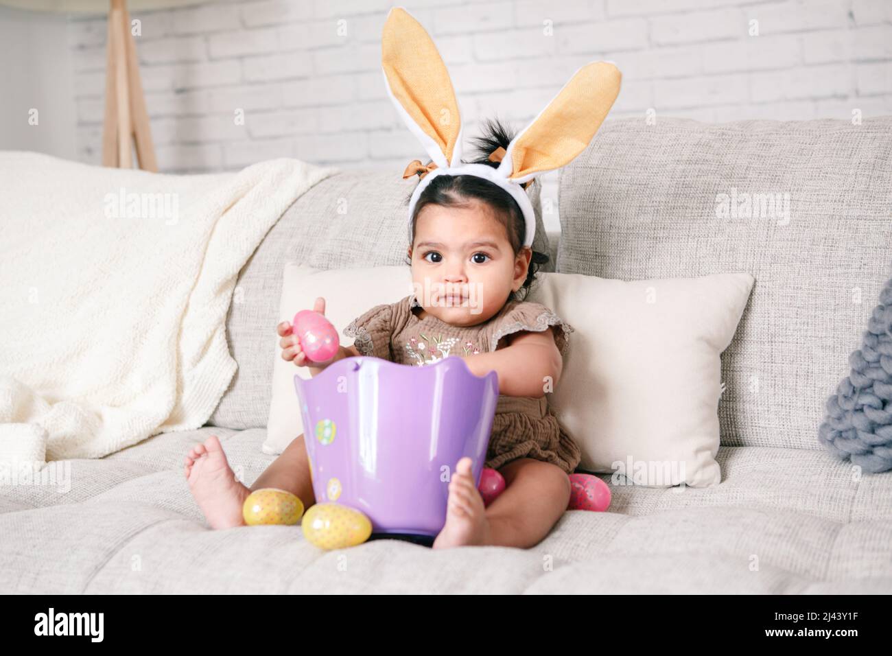 Cute Indian baby girl with pink bunny ears and basket of colorful eggs celebrating Easter holiday. Stock Photo