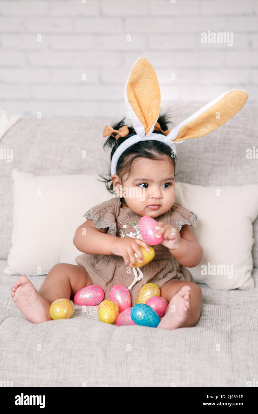 Cute Indian baby girl with pink bunny ears playing with colorful eggs candies toys celebrating Easter holiday. Stock Photo