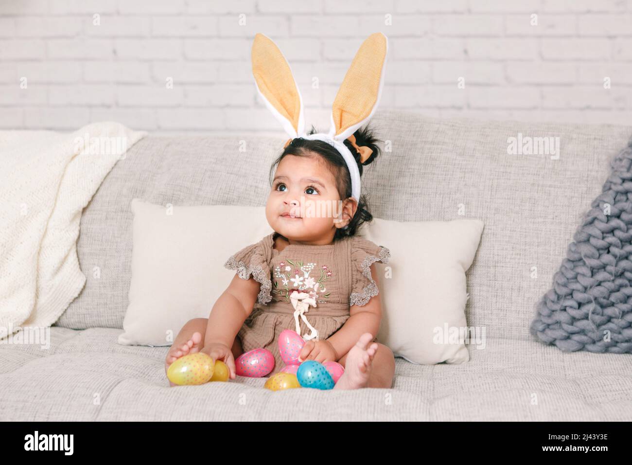 Cute Indian baby girl with pink bunny ears playing with colorful eggs candies toys celebrating Easter holiday. Stock Photo