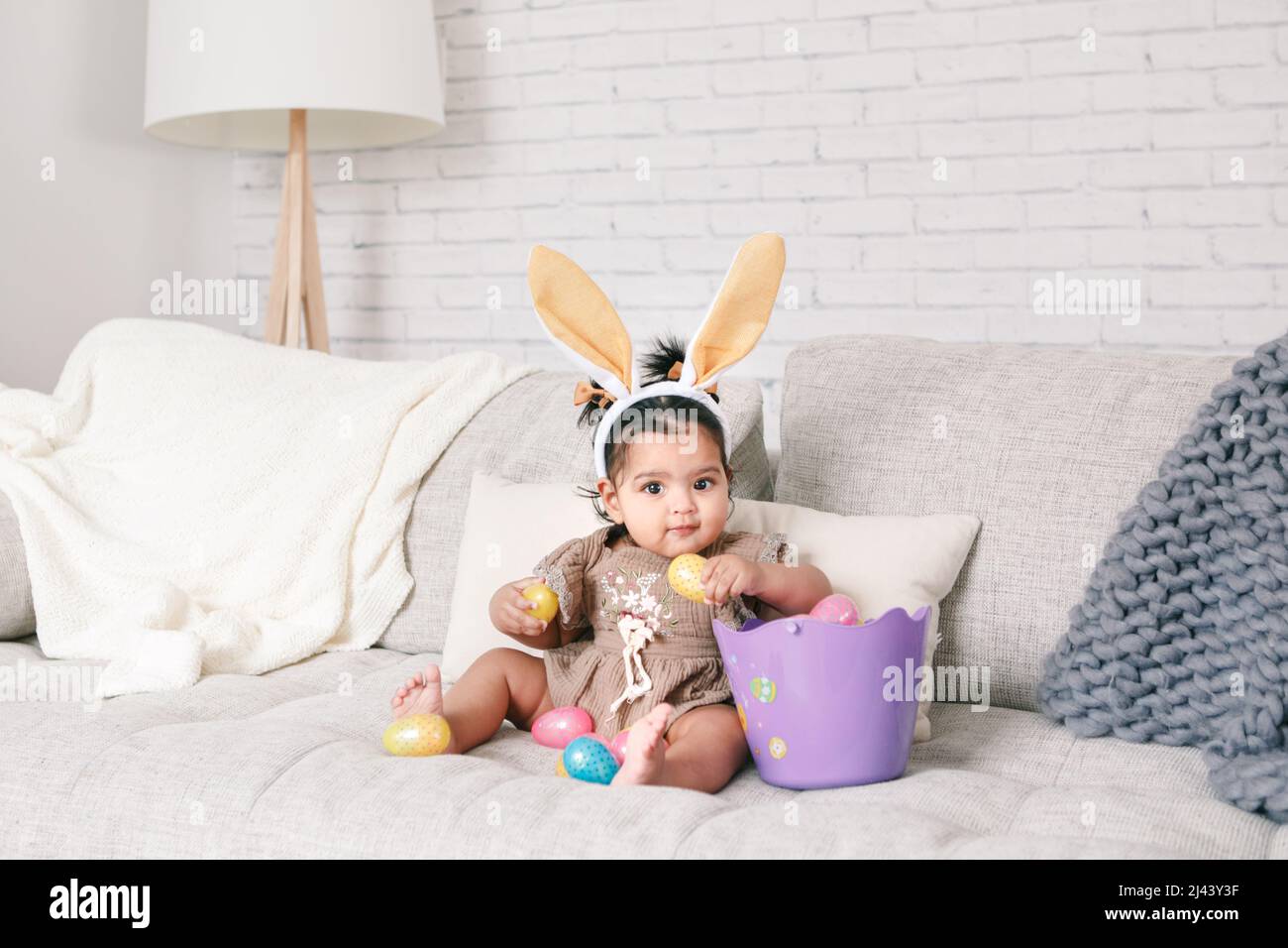 Cute Indian baby girl with pink bunny ears and basket of colorful eggs celebrating Easter holiday. Stock Photo