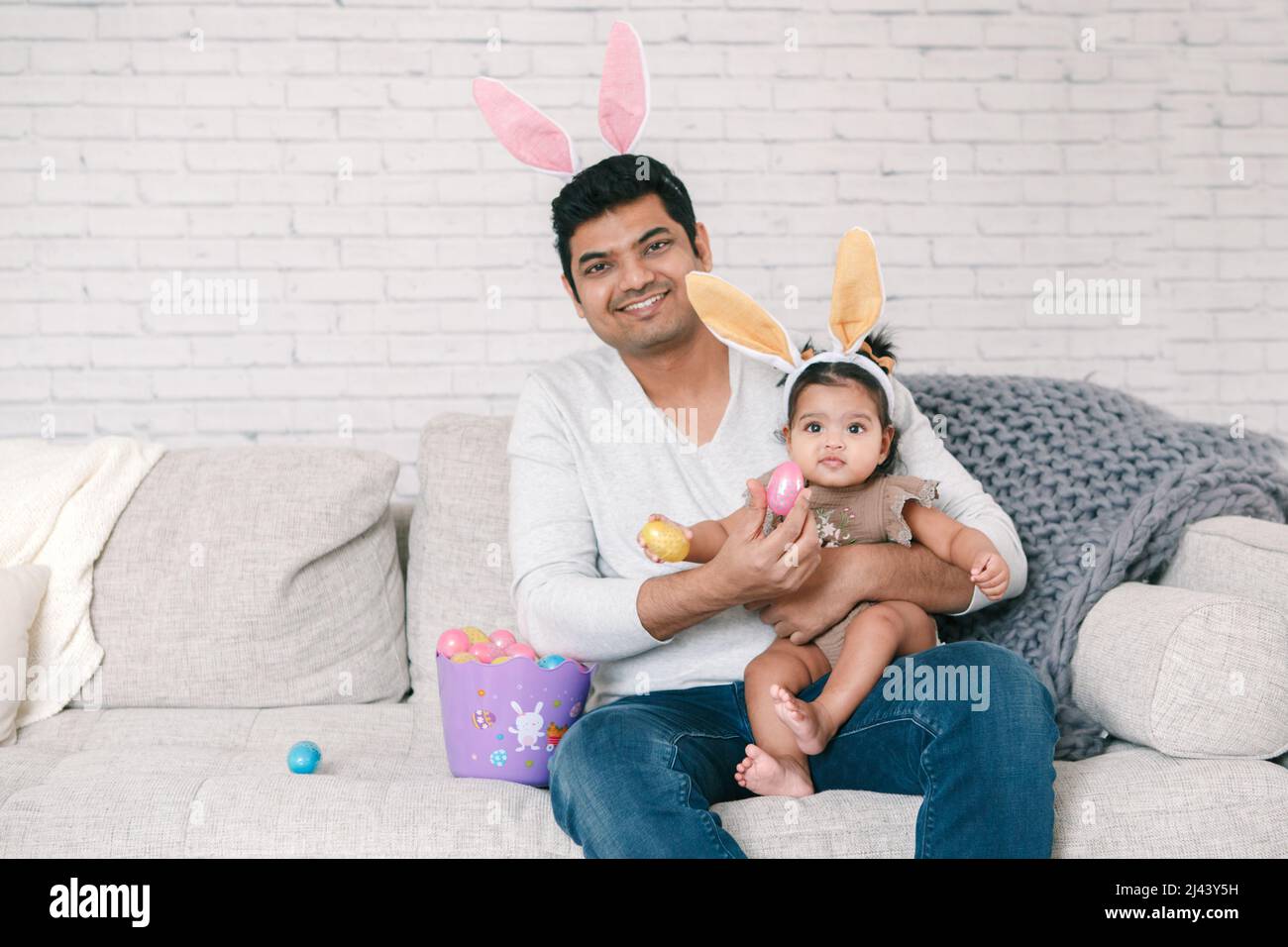 Indian father with baby girl daughter in bunny ears playing toy eggs in basket celebrating Easter. Stock Photo