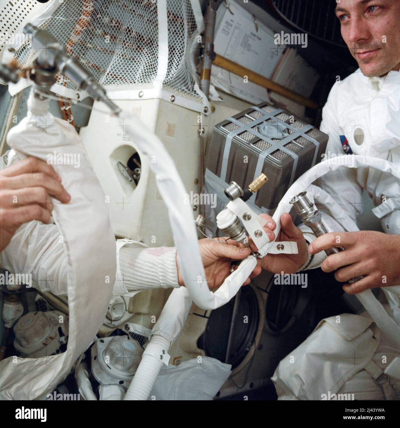 An interior view of the Apollo 13 Lunar Module (LM) during the trouble-plagued journey back to Earth. This photograph shows some of the temporary hose connections and apparatus which were necessary when the three Apollo astronauts moved from the Command Module (CM) to use the LM as a 'lifeboat'. Astronaut John L. Swigert Jr., command module pilot, is on the right. An unseen Lovell on the left holds in his right hand the feed water bag from the Portable Life Support System (PLSS). It is connected to a hose (center) from the Lunar Topographic (Hycon) Camera. In the background is the 'mail box,' Stock Photo