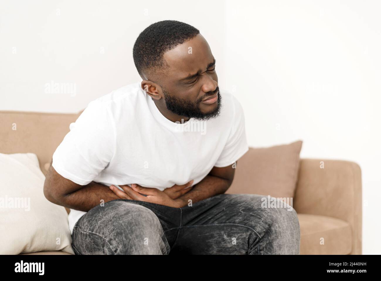Black man with abdominal pain suffering from painful abdominal cramp in stomach sitting on couch at home. Abdominal pain, stomach inflammation and app Stock Photo