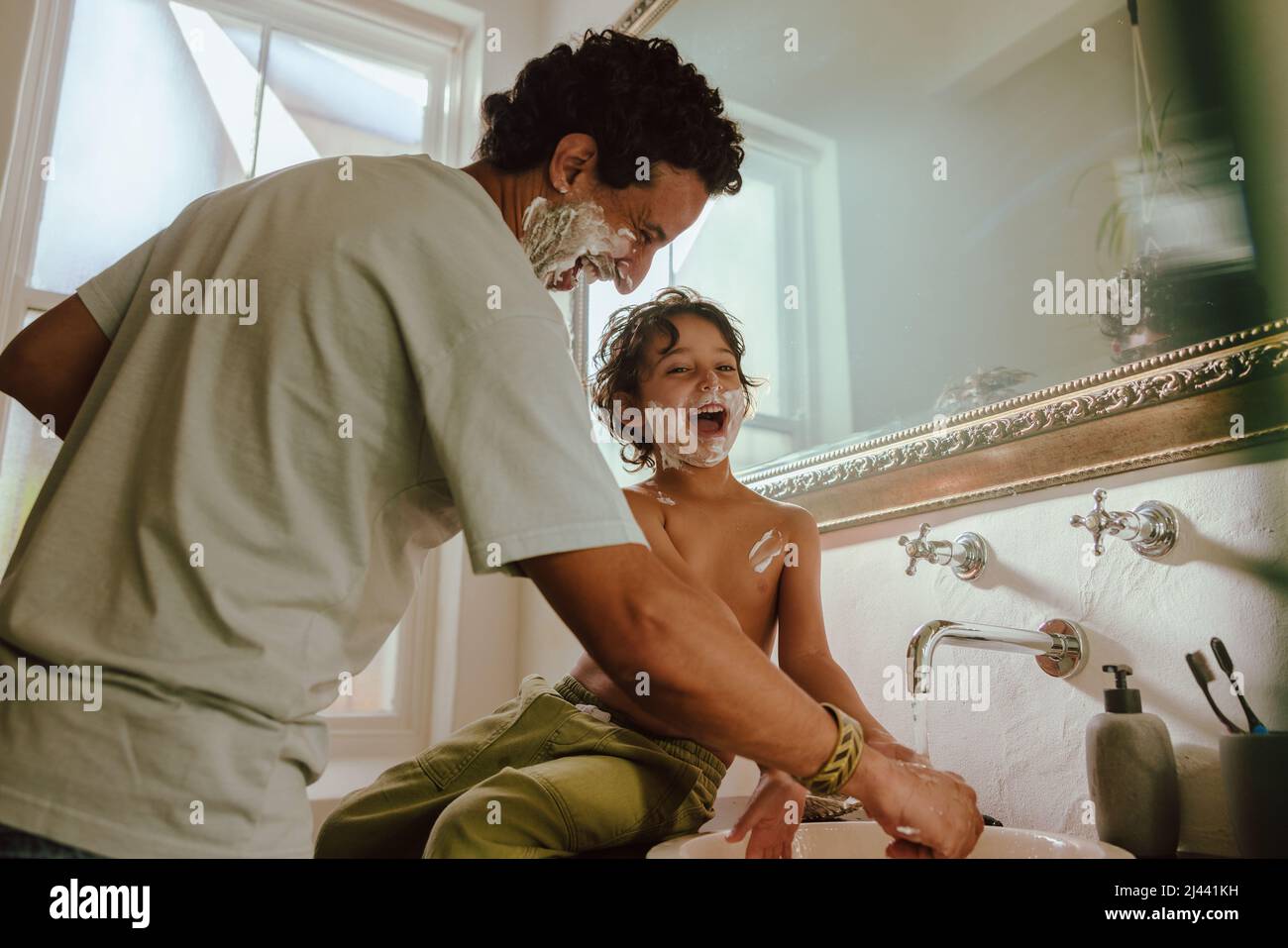 Cheerful father and son having fun with shaving foam in the bathroom. Father and son laughing happily with shaving cream on their faces. Loving father Stock Photo