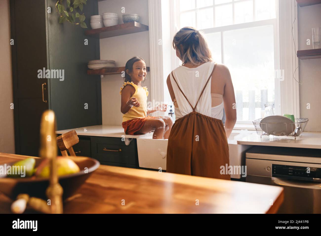 Single mother washing the dishes with her daughter at home. Happy little girl smiling at her mom while sitting on the sink countertop. Mother and daug Stock Photo