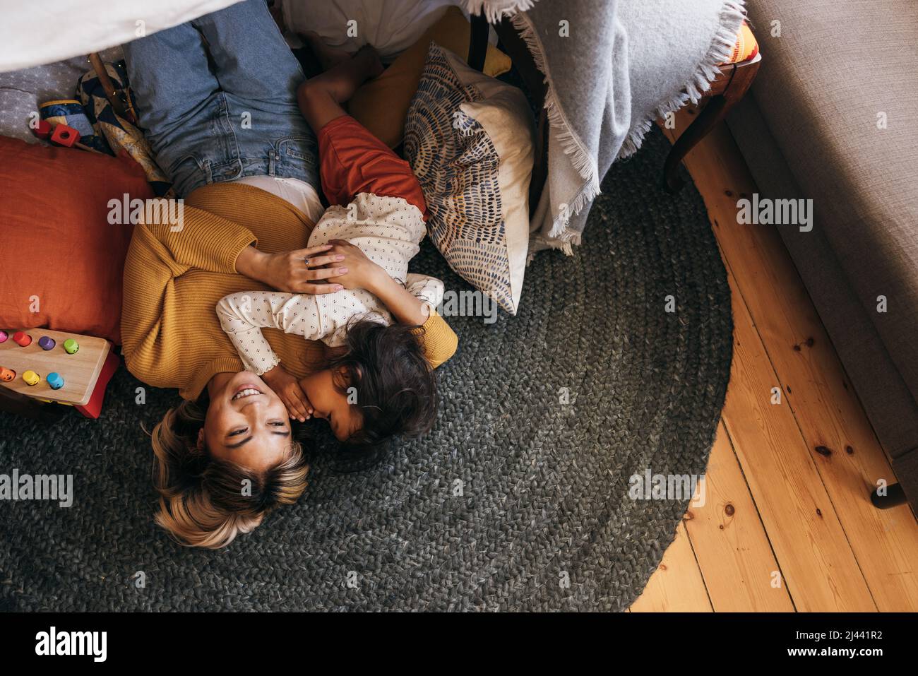 Top view of a young girl whispering into her mother's ear playfully. Cheerful mother and daughter lying on the floor in their play area. Mother and da Stock Photo