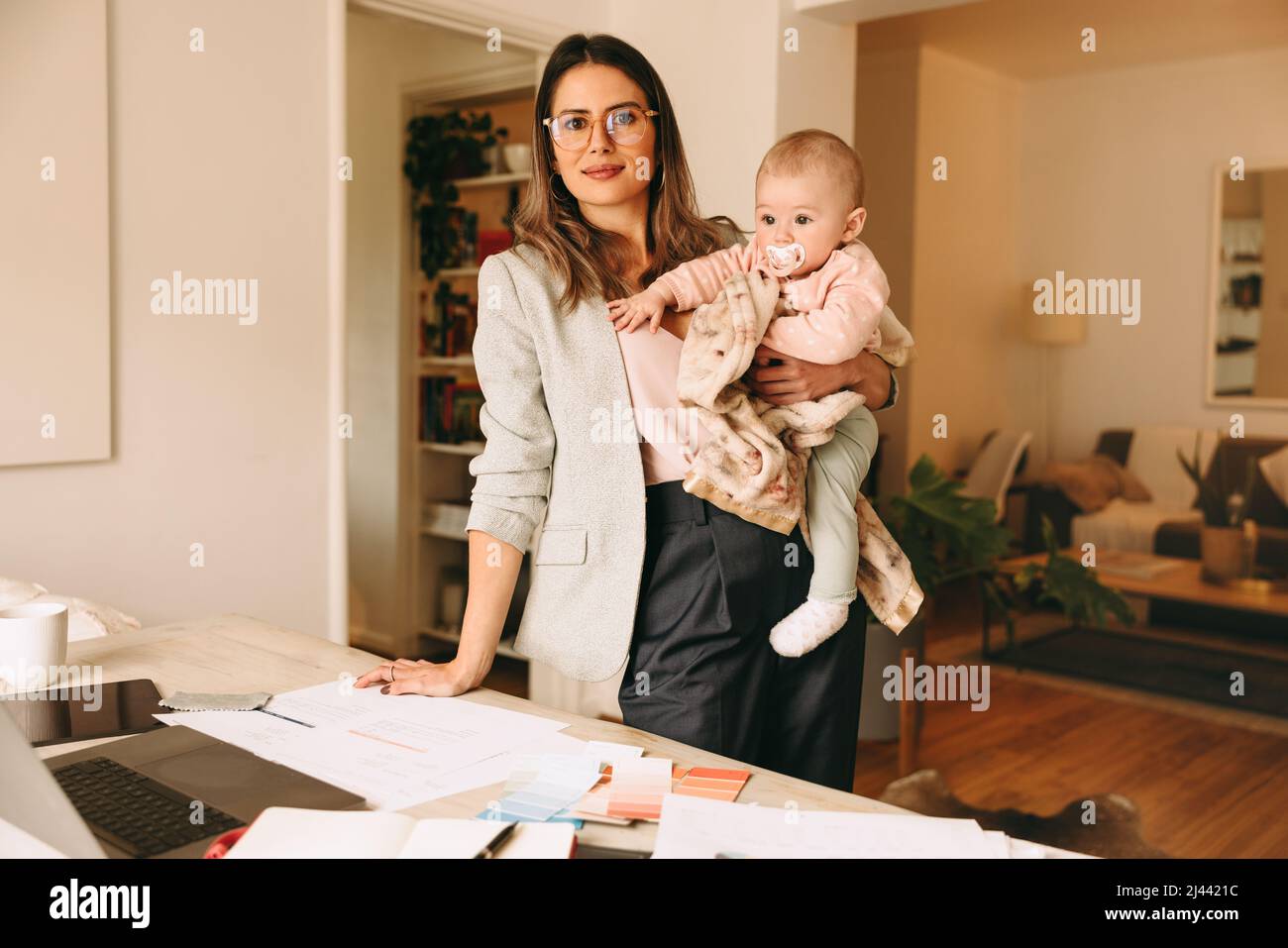 Creative businesswoman holding her baby while standing behind her desk. Working mom planning a new project in her home office. Female interior designe Stock Photo