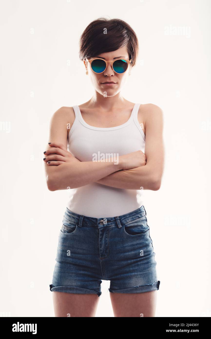 Trendy modern young woman with circular sunglasses or shades standing with folded arms in skimpy denim shorts staring at the camera Stock Photo