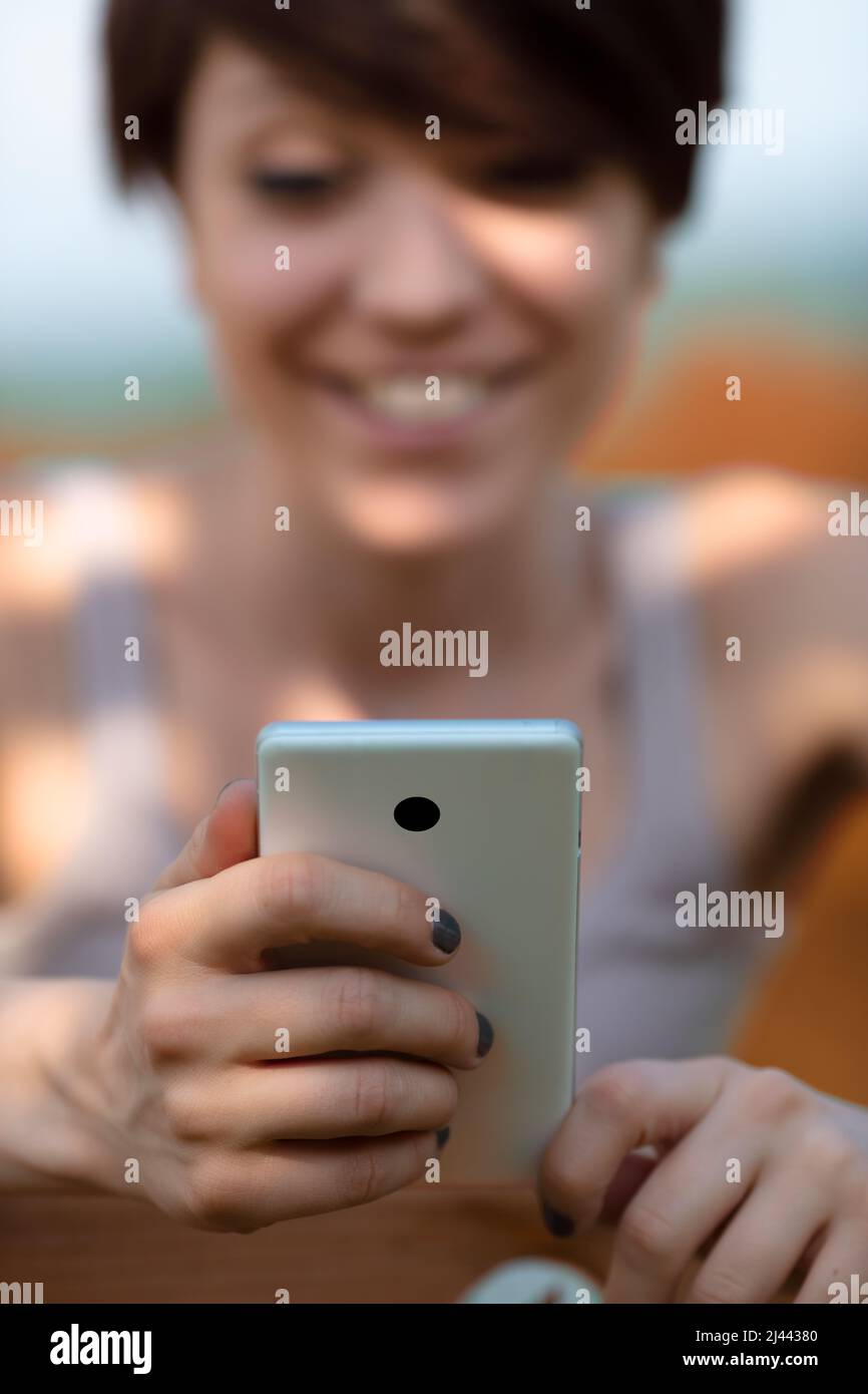 Young woman reading a message on her mobile with a smile in a close view with selective focus of her hand and the smartphone outdoors in dappled light Stock Photo