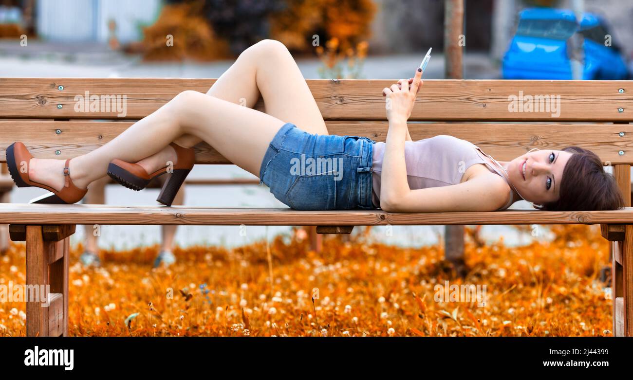 Pretty young woman relaxing lying on a park bench listening tomusic on her mobile phone turning to smile at the camera Stock Photo
