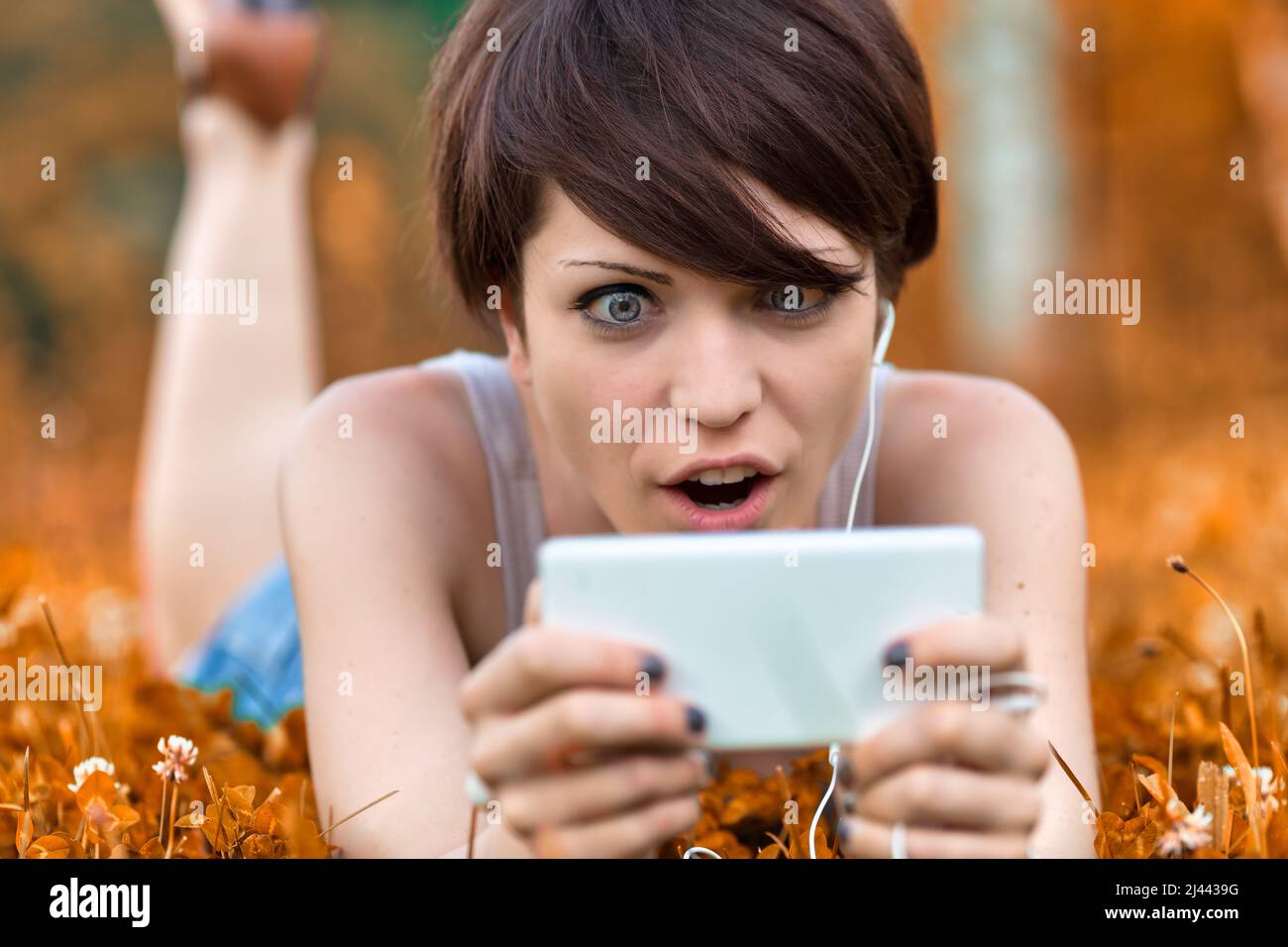 Young woman staring at her mobile in shock or amazement with wide staring eyes and open mouth as she lies outdoors in a meadow relaxing amongst wildfl Stock Photo