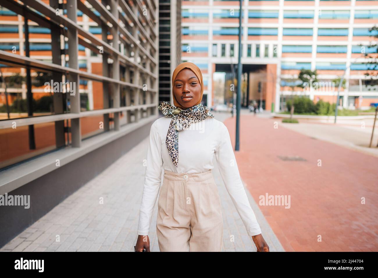 Confident African American female in modern outfit and Muslim hijab standing against contemporary skyscrapers of business center and looking at camera Stock Photo