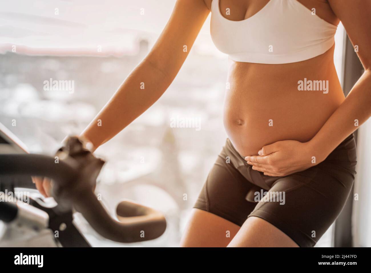 Pregnant woman training cardio on exercise spin bike at indoor fitness gym or home training on stationary bicycle. Closeup of fit girl holding Stock Photo