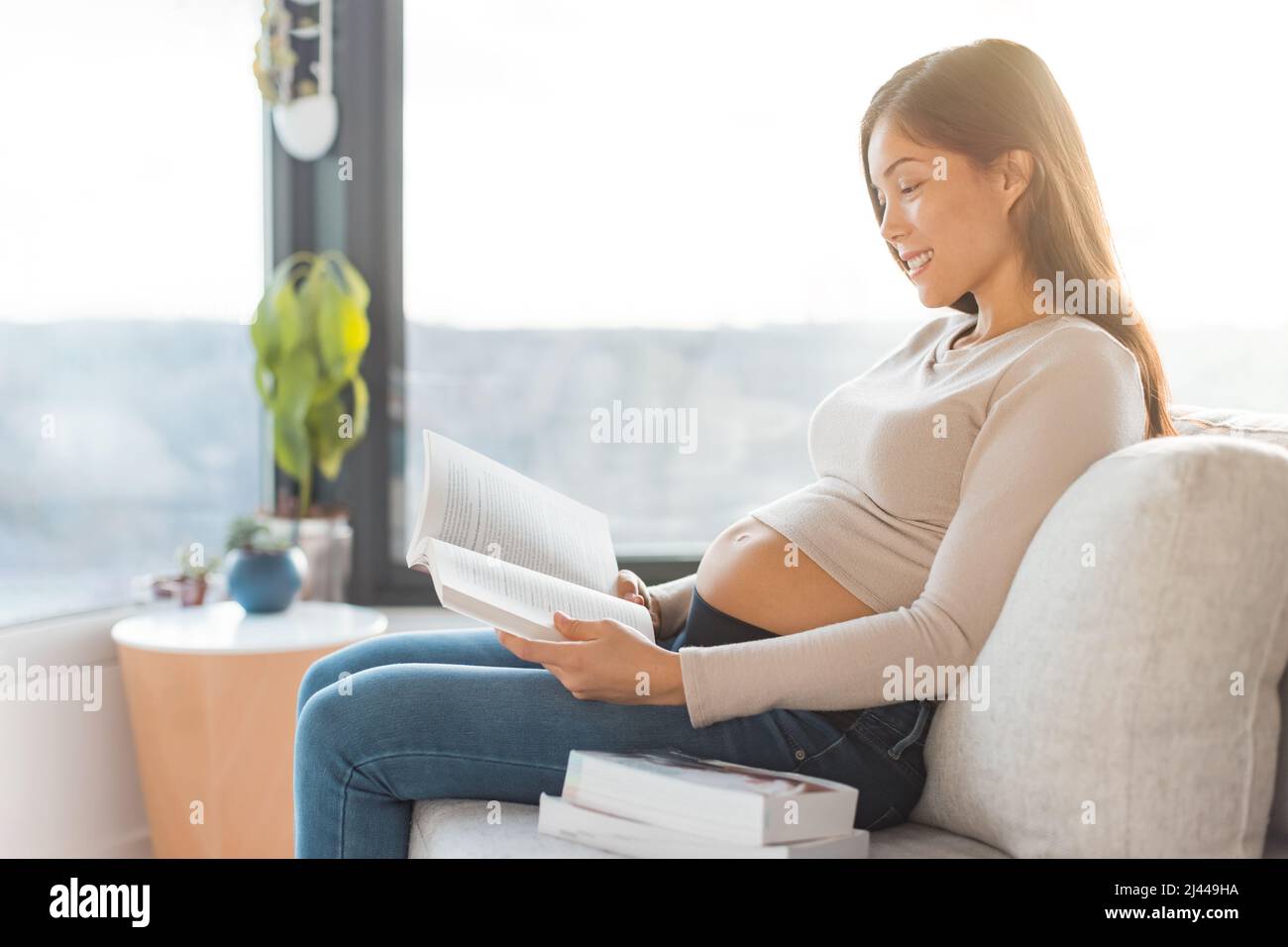 Pregnant Asian woman reading prenatal books preparing for labor, birth, newborn, sitting on sofa at home. Pregnancy lifestyle Stock Photo