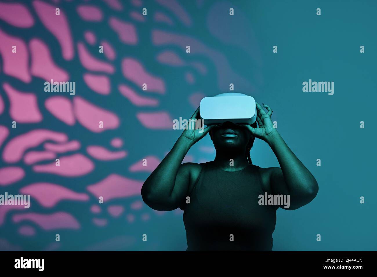 Black young woman in wireless goggles watching virtual reality against the pink pattern background Stock Photo