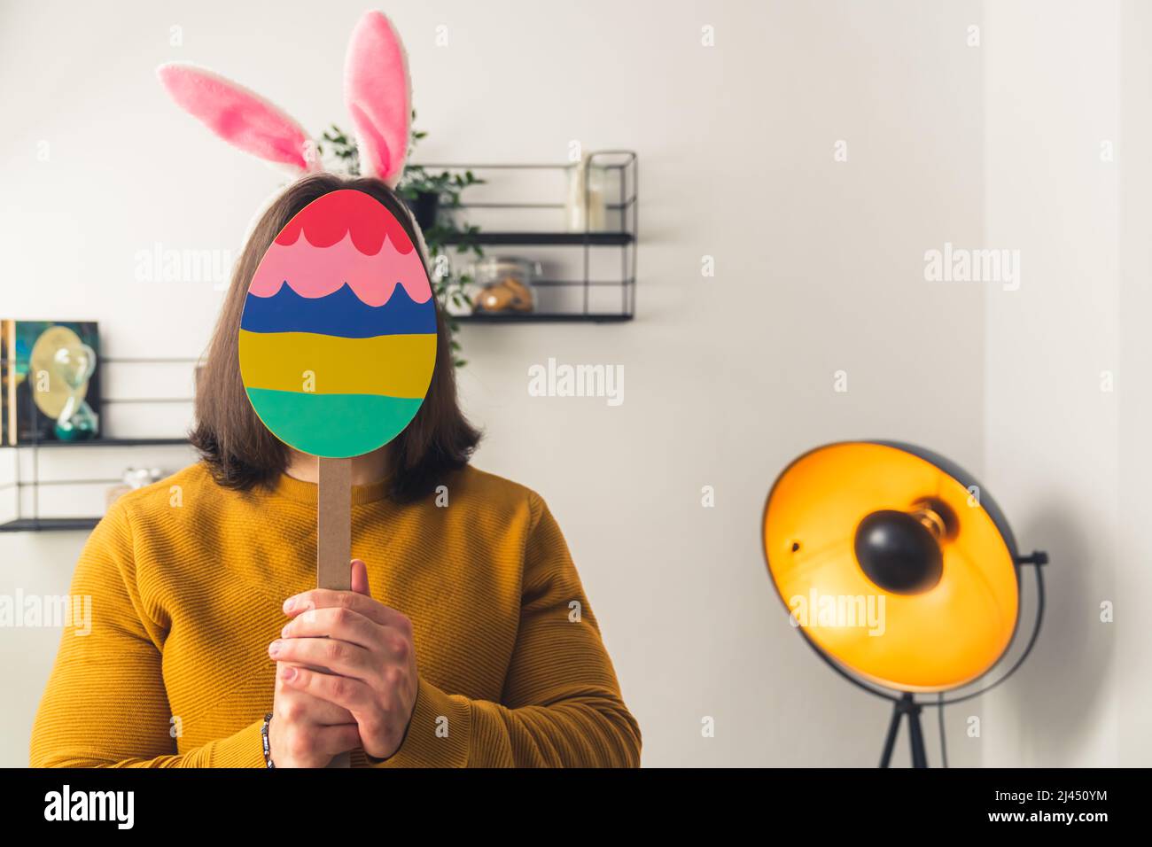 Young dark-haired man wearing fake rabbit ears hides face behind the paper Easter egg figure on a stick Easter concept medium closeup copy space home background . High quality photo Stock Photo