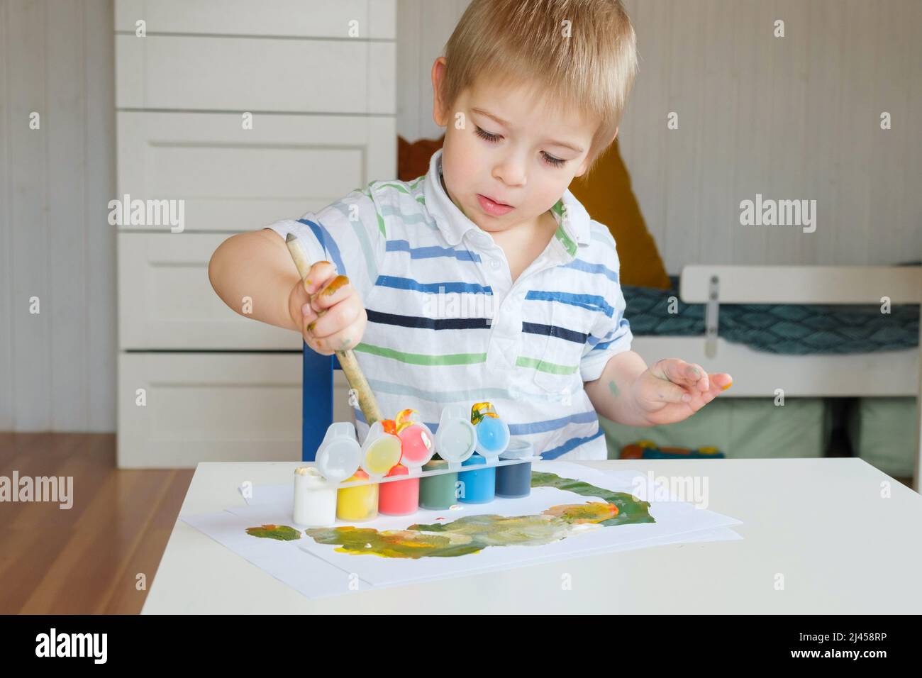 Little Boy Draws Picture On Paper. Preschool Child in striped clothes drawing with Paints at the Table at Home. Day. Real People. Creative little Kid. Stock Photo