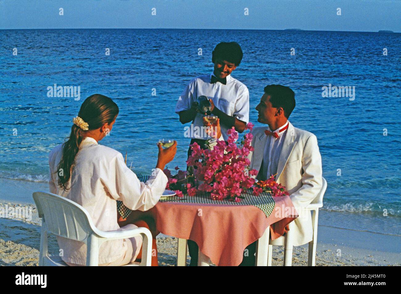 Maldives. Young couple in evening clothes being served dinner on the beach. Stock Photo