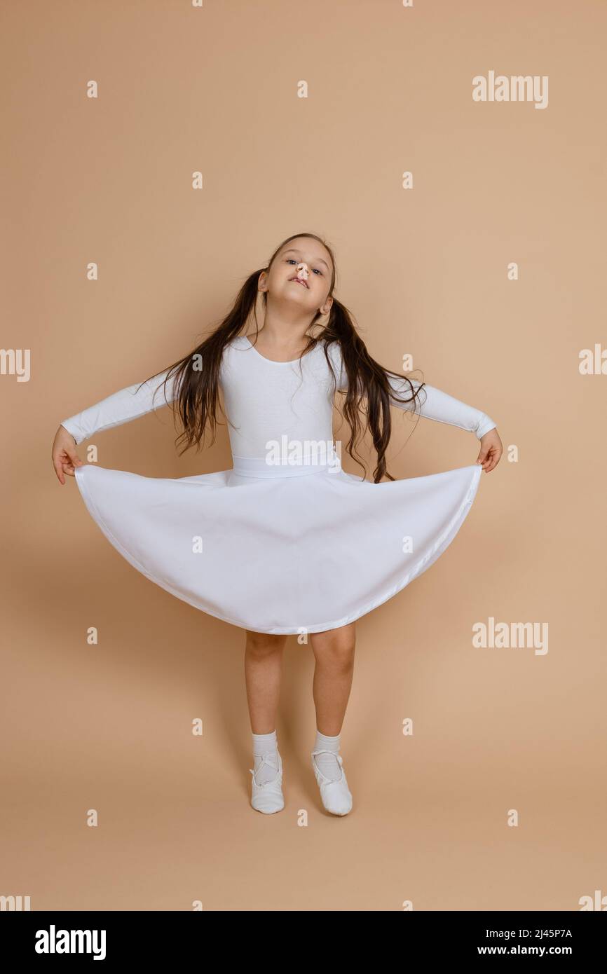 Young cute smiling girl with long dark hair in white dress, socks and gymnastics shoes standing, holding dress with hands, tip-toeing, having fun on Stock Photo