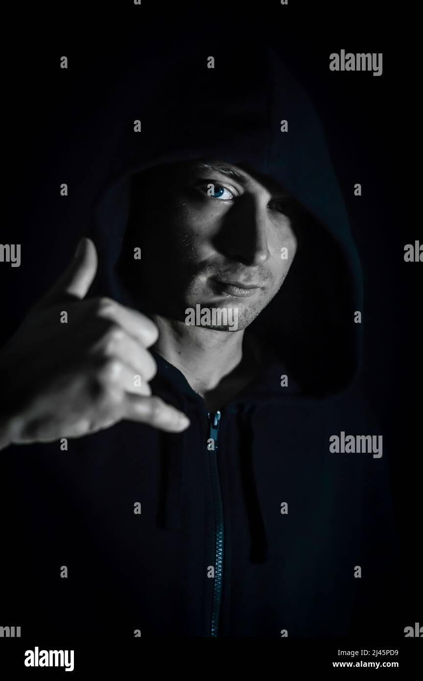 Shaka - Portrait of a young hooded man who in the shade makes a sign of relaxation with his hand, hang loose, salutation Stock Photo