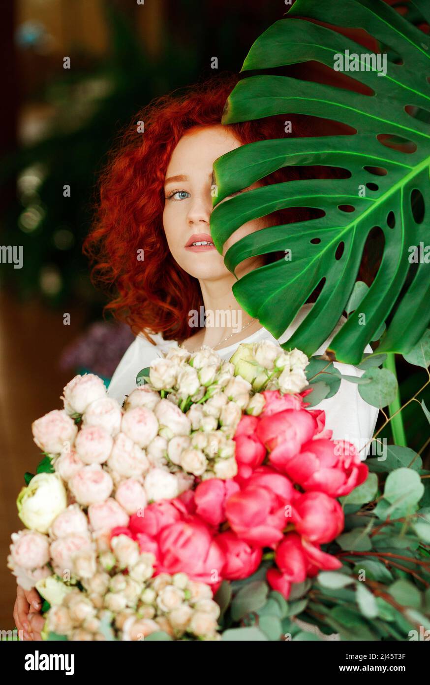 Portrait of a beautiful red-haired curly girl with a bouquet of spring flowers in her hands, hiding part of her face behind a green leaf with a flower Stock Photo
