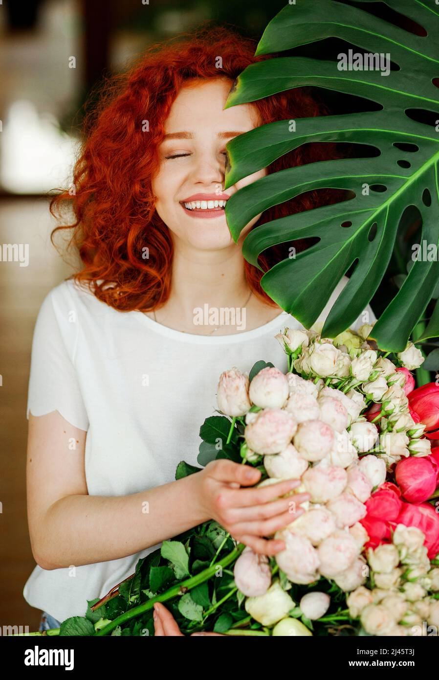 Portrait of a beautiful red-haired curly girl with a bouquet of spring flowers in her hands, hiding part of her face behind a green leaf with a flower Stock Photo