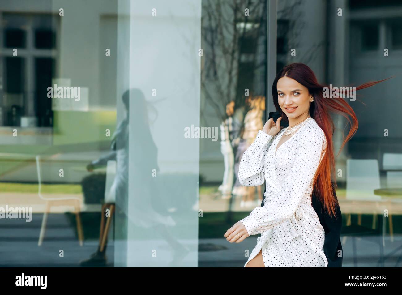Stylish red-haired woman in a white dress walking down the street near ...
