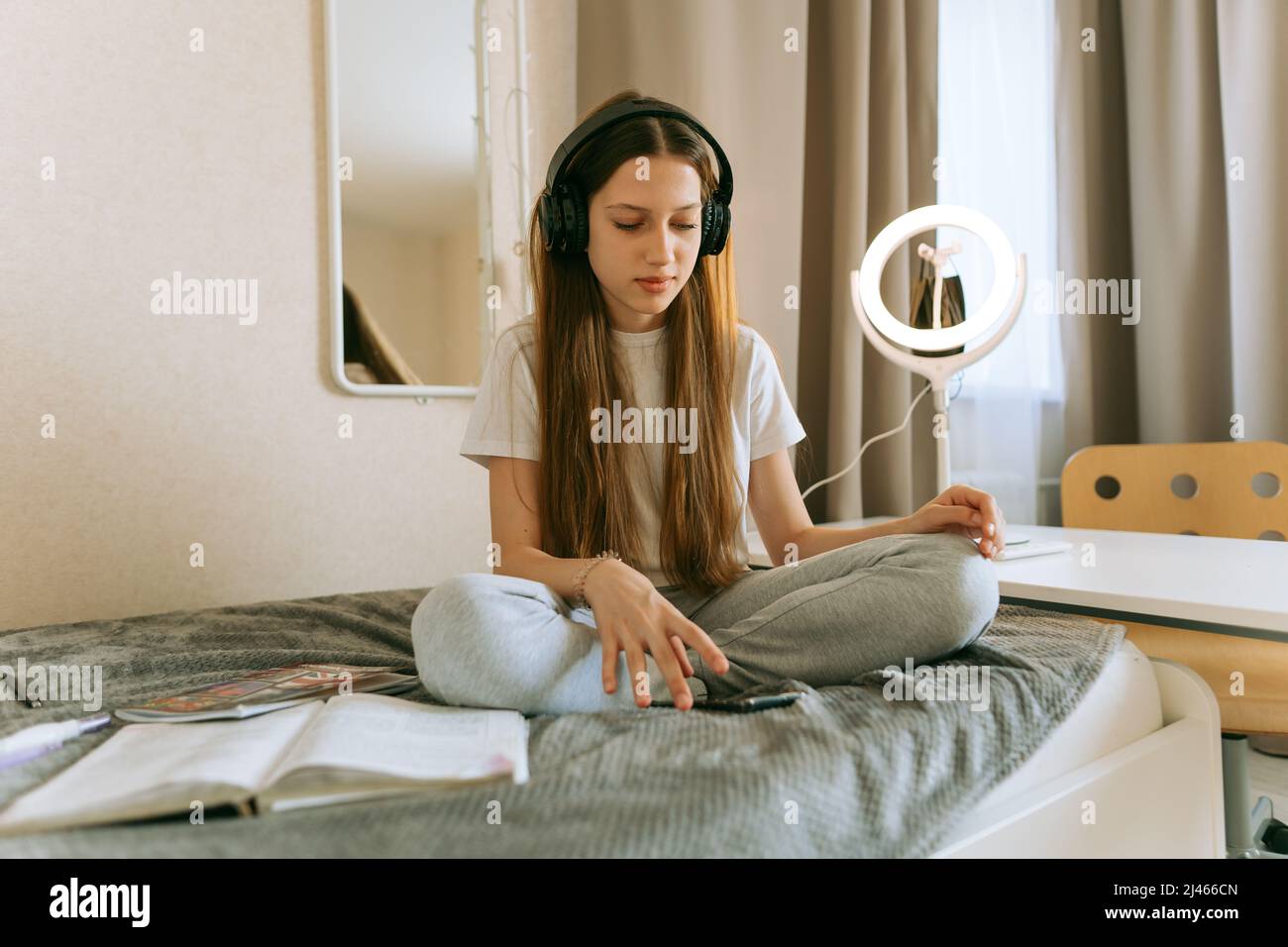 Young teenage girl in headphones and with smartphone is doing homework sitting on bed. A girl of generation Z with long hair, she is studying intently Stock Photo