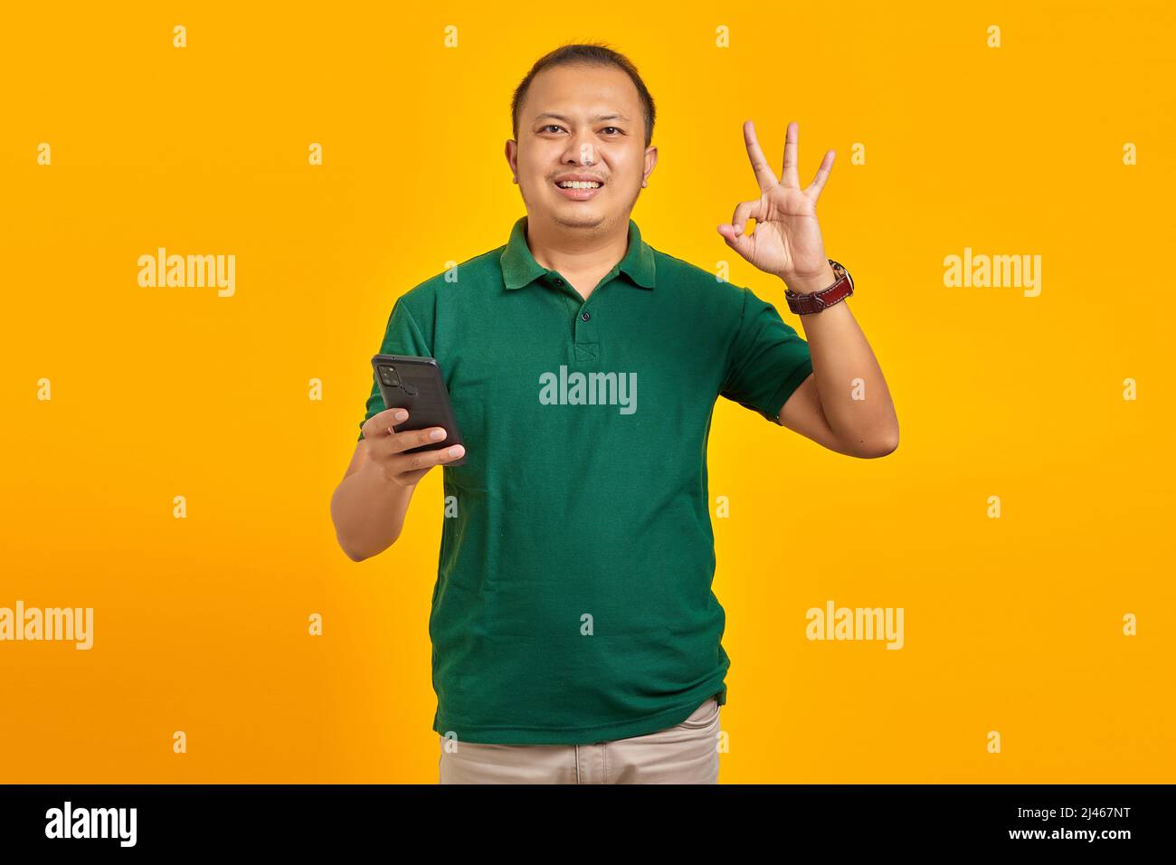 Smiling young man using smartphone and doing okay gesture on yellow background Stock Photo