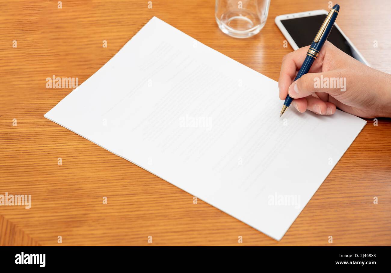 Man holding a pen on a blank white paper, close up above view, wooden office table background. Male hand signing on empty document, copy space Stock Photo