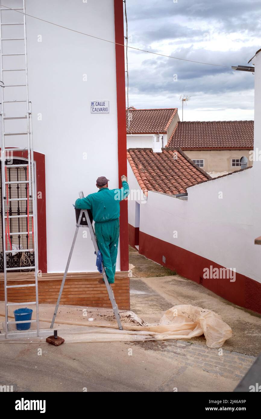 A painter and decorator paints a house in the centre of Berrocal, Huelva, Spain. He wears green overalls and stands on a ladder Stock Photo