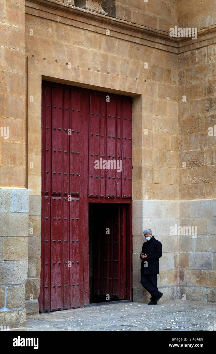 A man wearing a covid mask stands outside a large red wooden door at Salamanca Cathedral, Spain. He uses a mobile phone Stock Photo