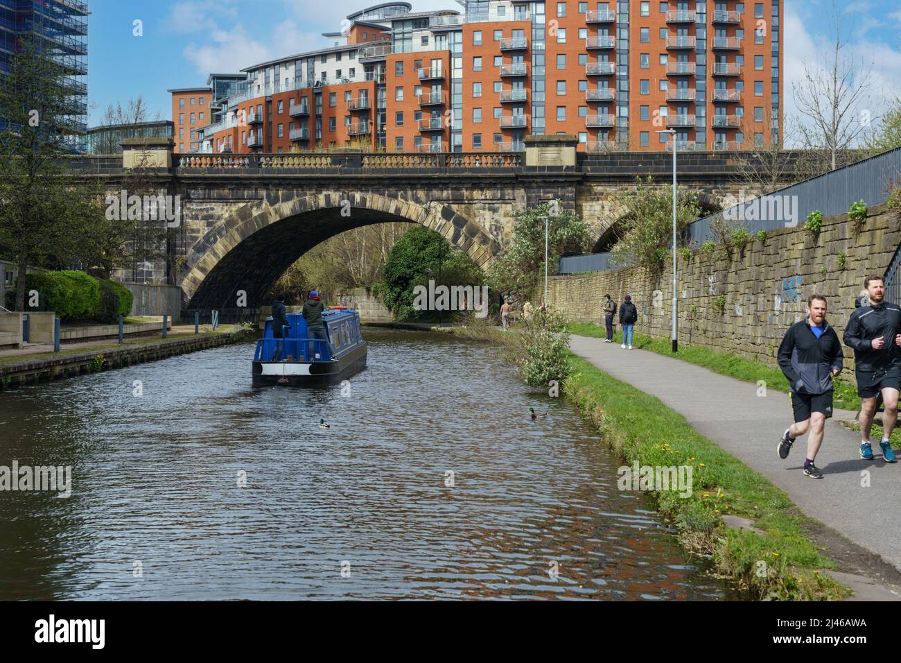 Two people travelling on a Barge along the Leeds and Liverpool Canal with two joggers running along the towpath, Leeds, West Yorkshire,UK. Stock Photo