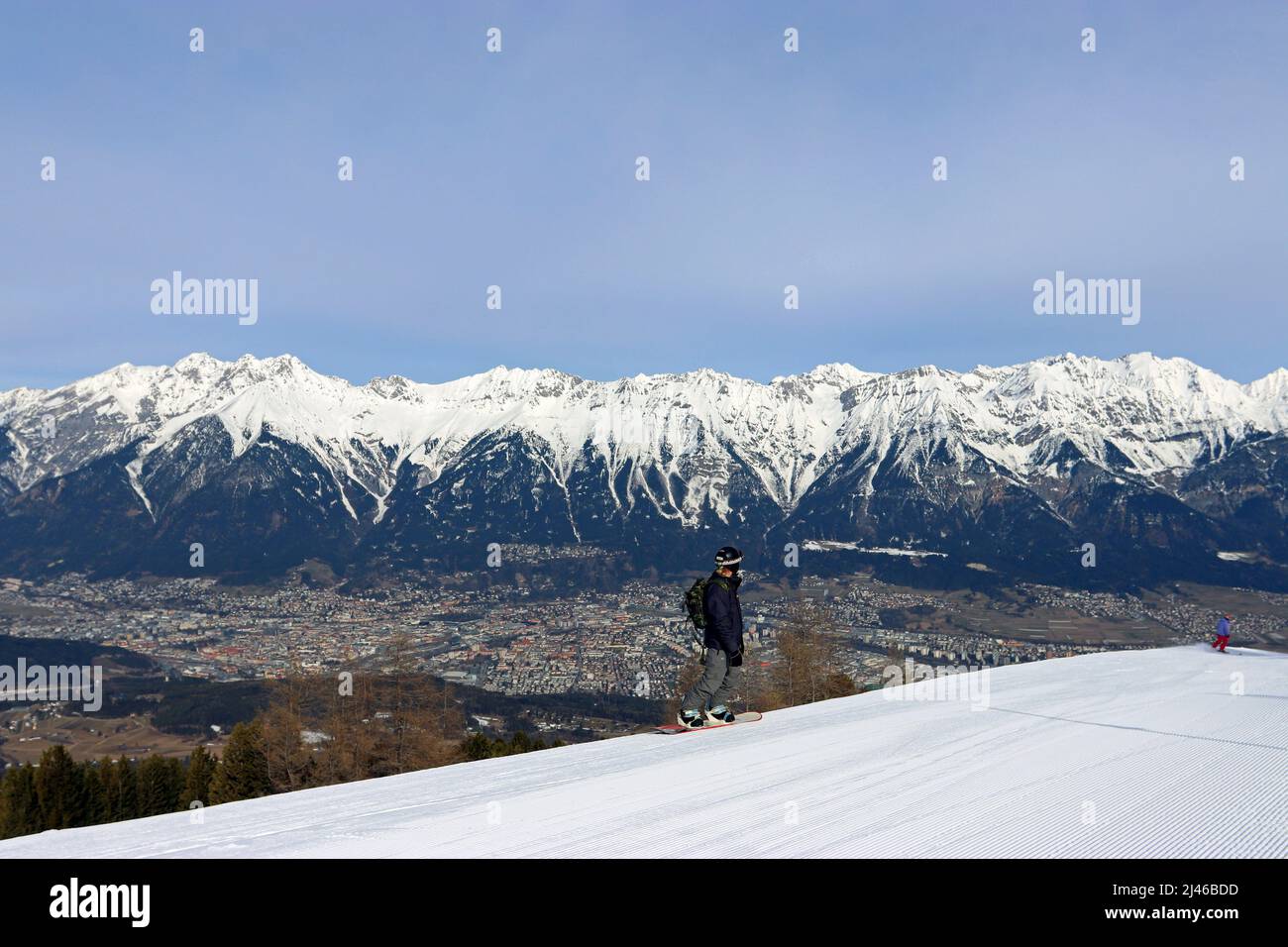 A snowboarder stands on Patscherkofel mountain. Innsbruck spreads out below against the snowy mountain backdrop of the Nordkette mountain range Stock Photo