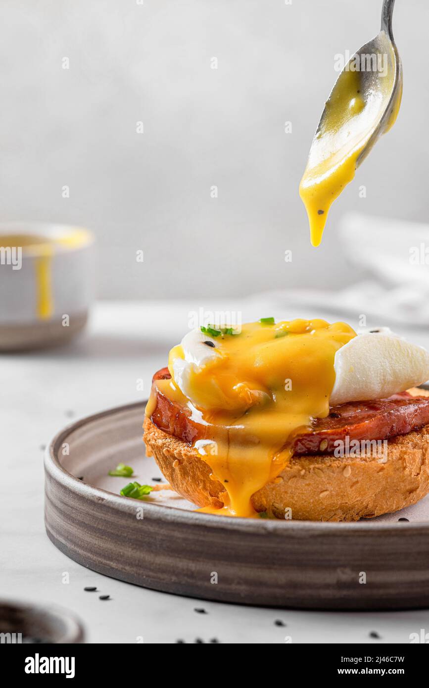 Pouring Hollandaise sauce in egg benedict with poached egg, ham, onion on slice toast for tasty breakfast. vertical orientation. English brunch Stock Photo