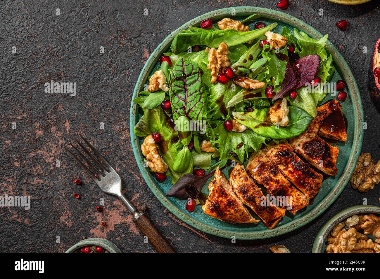 Grilled chicken breast fillet and mix salad with walnuts, pomegranate and balsamic sauce on dark background with fork. top view. healthy food Stock Photo